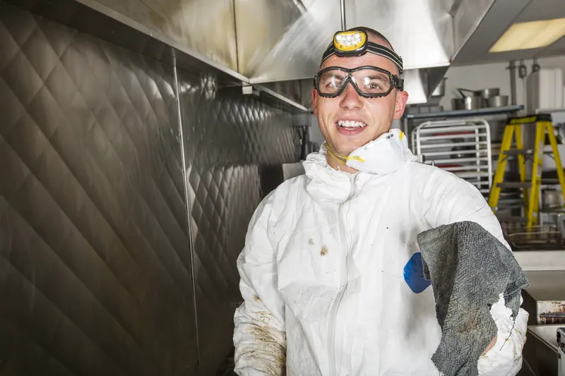 Technician in white coveralls holding a cleaning cloth beside the cook line in a commercial kitchen