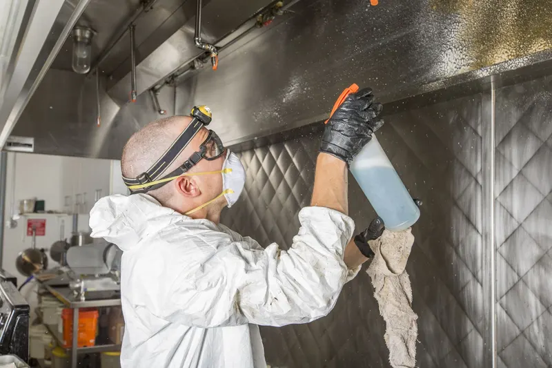 Technician in a respirator spraying degreaser up into a stainless exhaust canopy from below