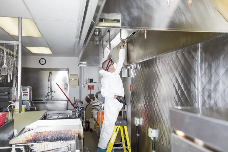 Hood cleaning technician on a yellow step ladder reaching into a stainless canopy in a full commercial kitchen